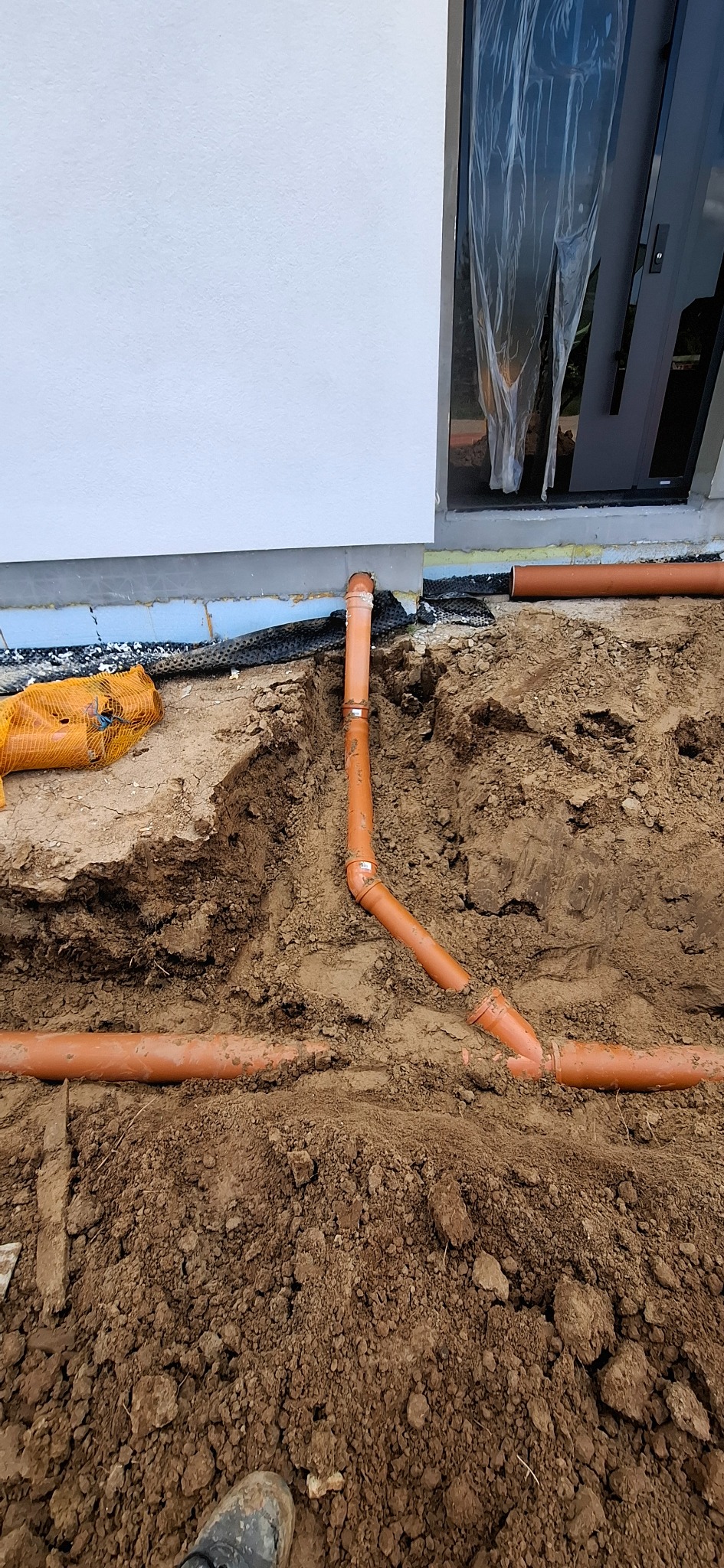 Brązowe rury kanalizacyjne ułożone w ziemi przy ścianie budynku z oknem. Widoczne złączenia rur i fragment buta w dolnym rogu kadru.