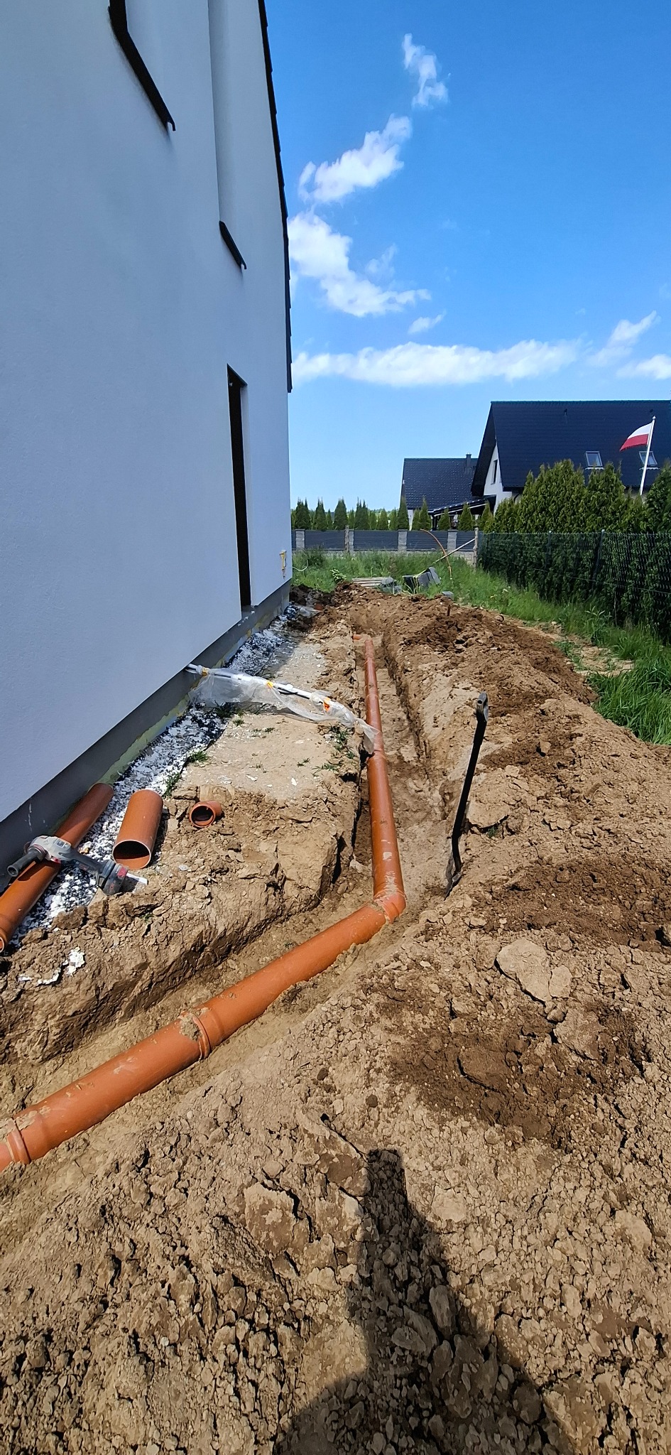 Wykonanie przyłącza kanalizacyjnego: pomarańczowe rury ułożone w wykopie obok budynku, widoczny fragment elewacji i niebieskie niebo w tle.
