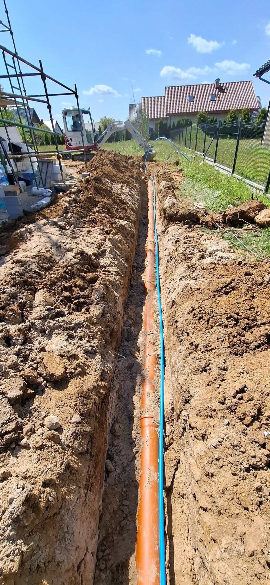 Wykop z pomarańczową rurą i niebieskim przewodem, widoczny mini-koparka i rusztowanie w tle, słoneczny dzień, instalacja w ziemi.