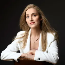 Portret kobiety w białym żakiecie, z blond włosami opadającymi na ramiona, siedzącej na krześle z rękami skrzyżowanymi na oparciu, na ciemnym tle.