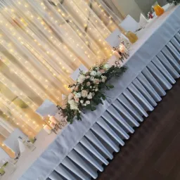 Uroczysty stół weselny z białym obrusem, falbaną, kwiatową dekoracją, świecami i napojami, oświetlony girlandą świetlną w tle.