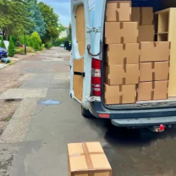 Otwarty tył białego busa zapakowanego kartonami z napisem, dwa kartony na chodniku, ulica w tle.