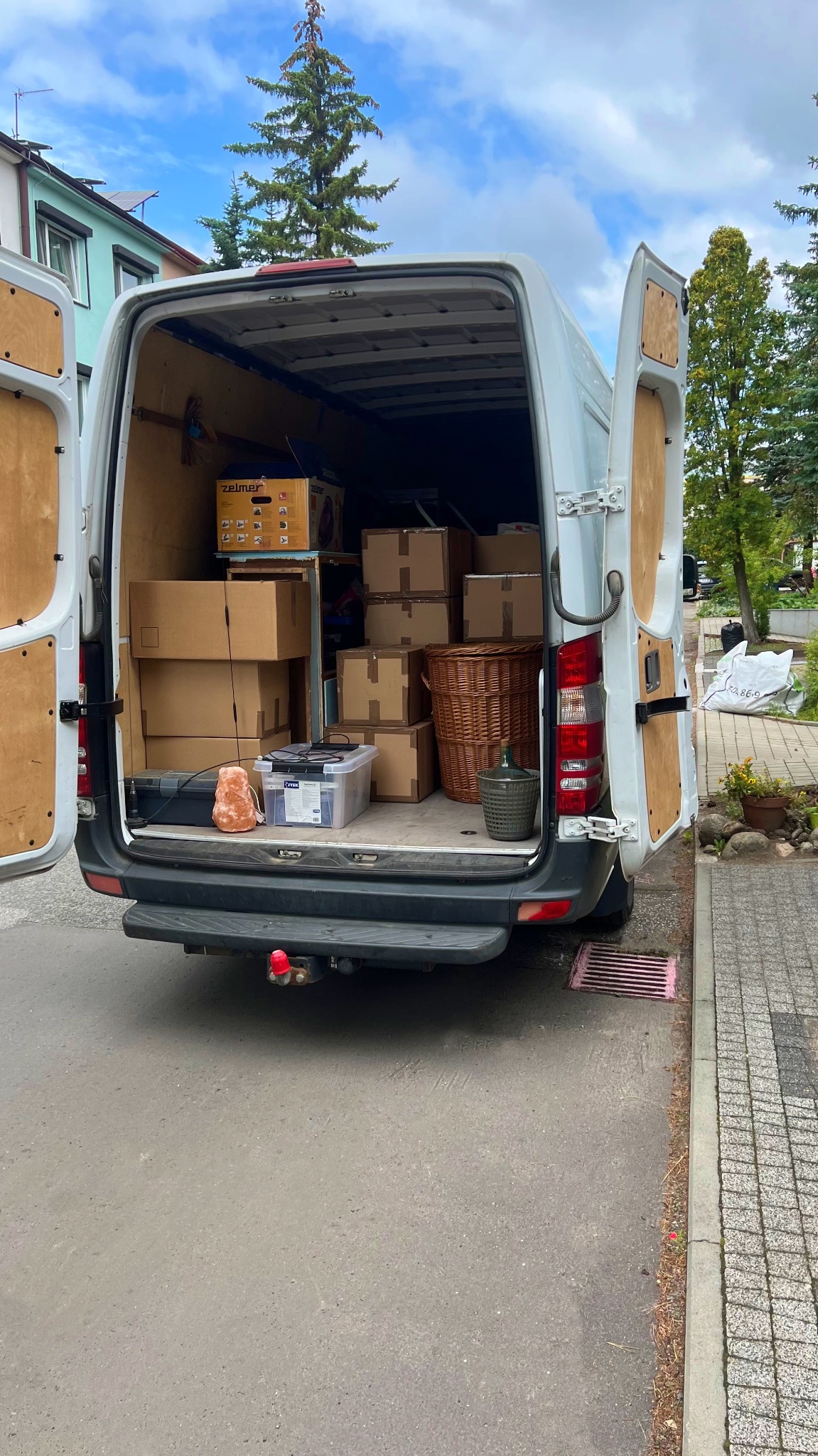 Otwarty tył białego busa wypełnionego kartonami, koszem wiklinowym, lampą solną i innymi przedmiotami podczas załadunku na zewnątrz, widok z poziomu ulicy.