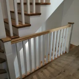 Schody drewniane z białymi balustradami i drewnianą poręczą, widok z dołu na fragment schodów z parkietem na podłodze.