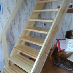 Drewniane schody prowadzące na antresolę, widok z dołu, jasne drewno, dekoracyjna tapeta w tle, fragment sofy widoczny po prawej stronie.