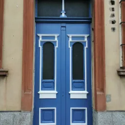 Dwuskrzydłowe, niebieskie drzwi wejściowe z białymi zdobieniami, osadzone w brązowej ramie na tle beżowej ściany z kamienną podstawą.