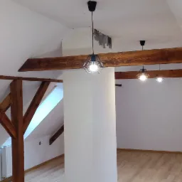Wnętrze poddasza z drewnianą konstrukcją stropu, białymi ścianami, jasną podłogą i trzema lampami wiszącymi w metalowych kloszach o geometrycznym kształcie, widoczne ślady po remoncie na suficie.