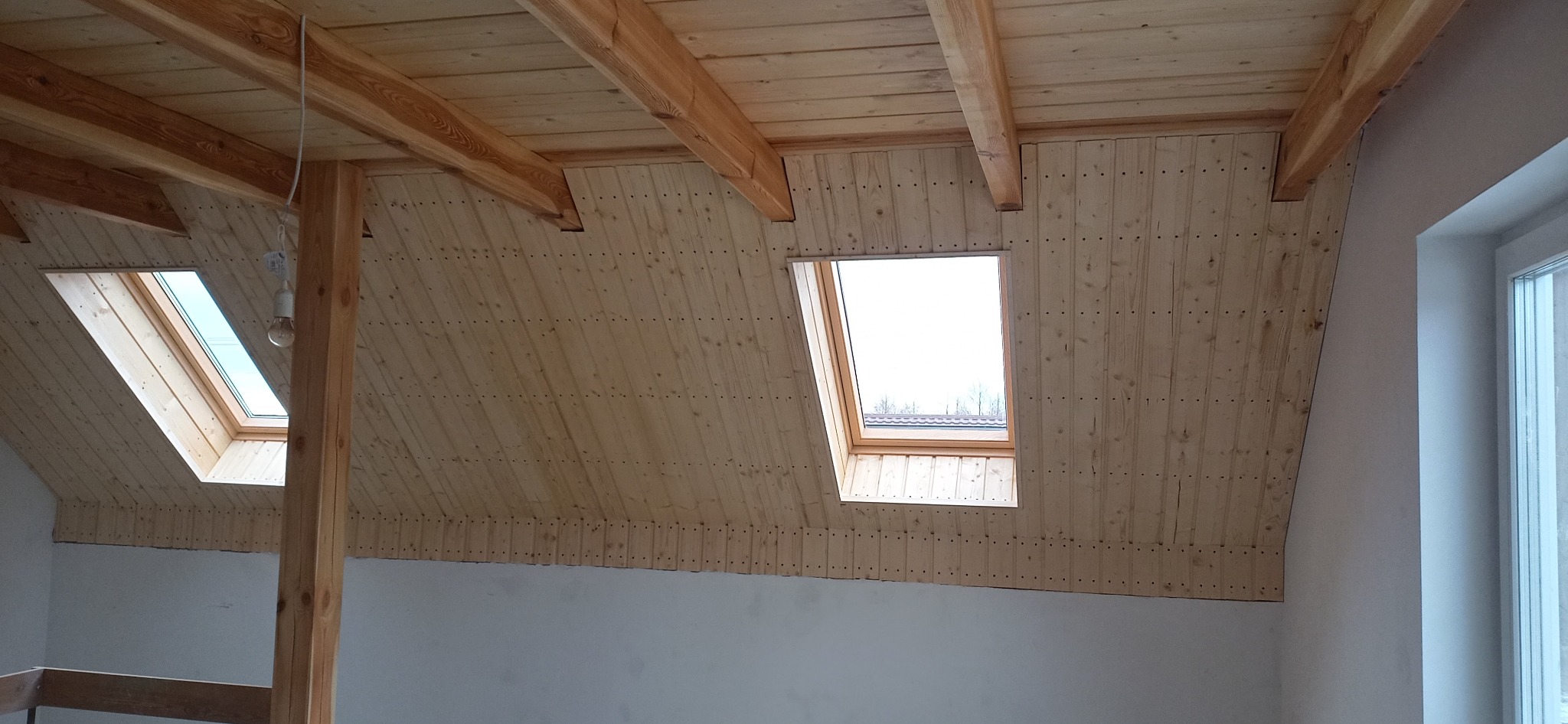 Wykończone poddasze z drewnianą boazerią, widoczne dwa okna dachowe i drewniane belki stropowe, surowa żarówka zwisająca z sufitu.
