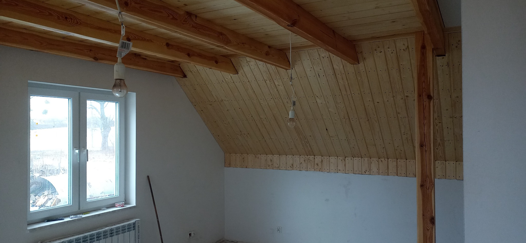 Montaż drewnianej boazerii na poddaszu w Chojnicach, widoczne belki stropowe i surowa ściana, okno z widokiem na zewnątrz.