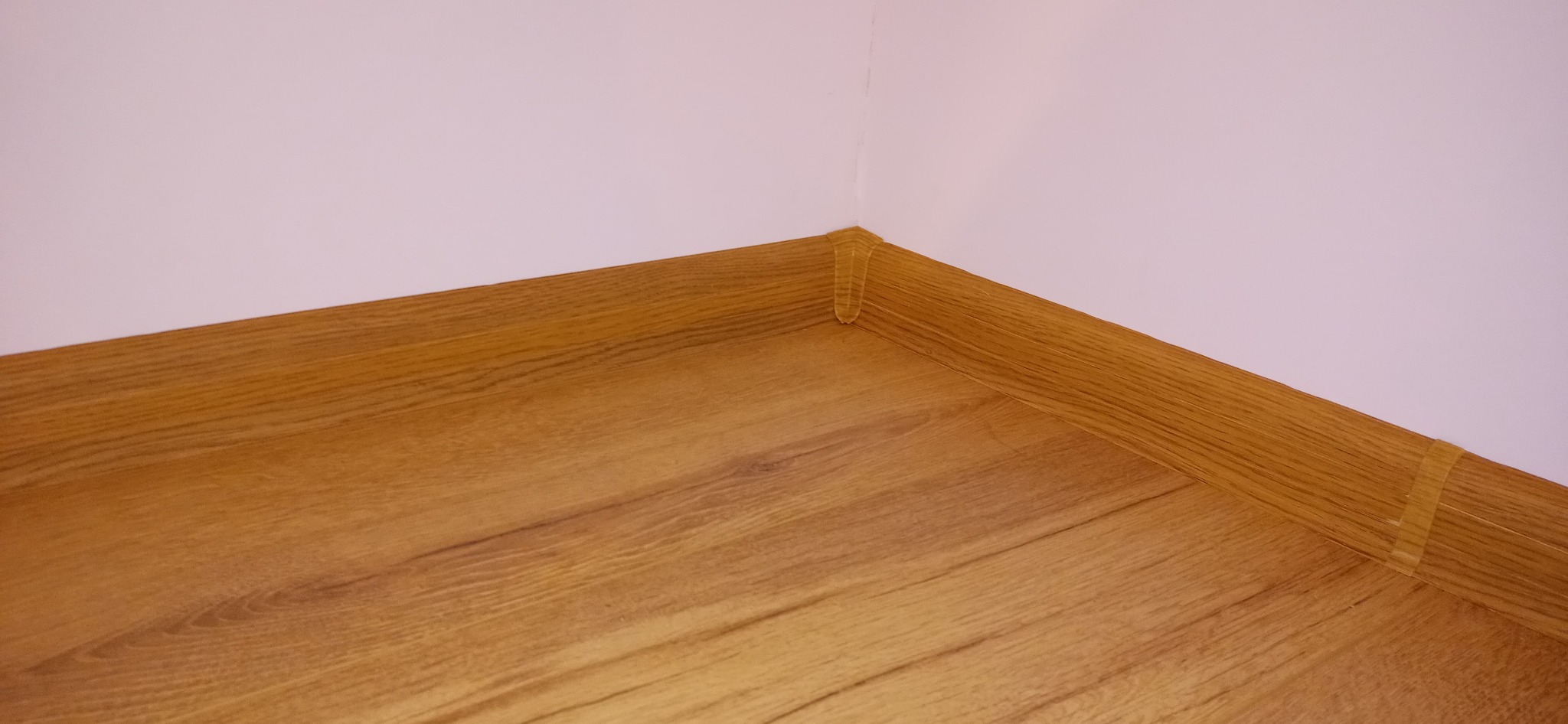 Szczegół wykończenia podłogi laminowanej z listwą przypodłogową przy ścianie, zabezpieczoną taśmą malarską w rogu pomieszczenia.