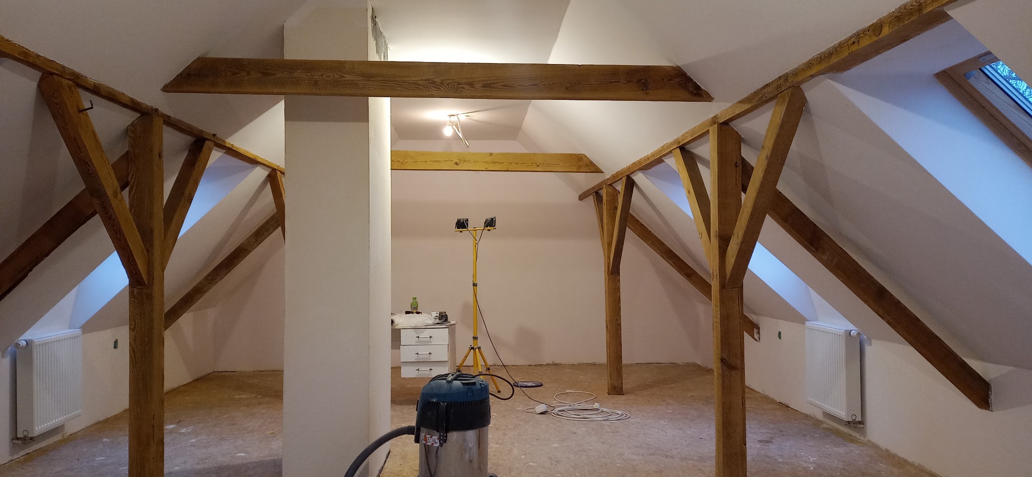 Poddasze w trakcie remontu, widoczne drewniane belki konstrukcyjne, białe ściany, grzejnik, lampa budowlana na statywie i odkurzacz przemysłowy na podłodze.