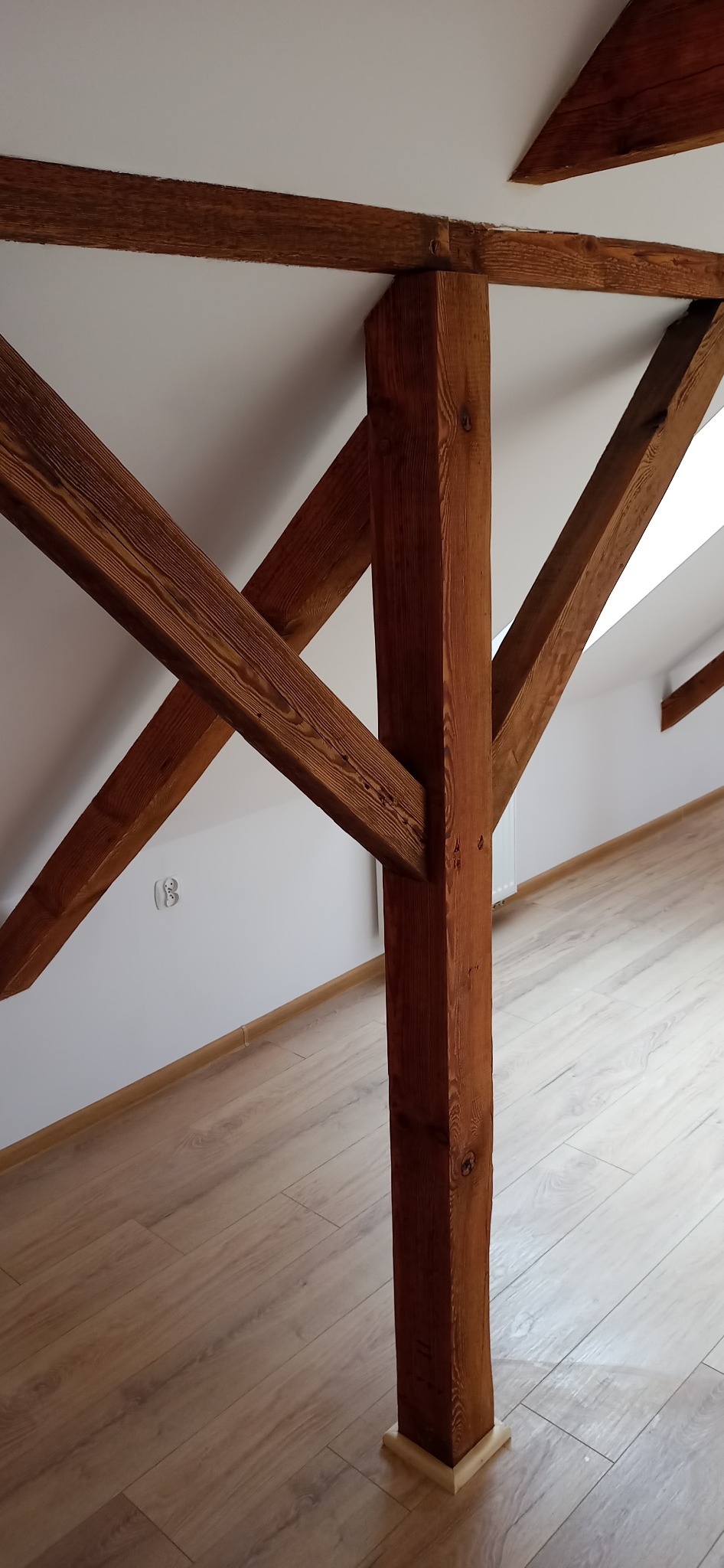 Drewniana konstrukcja stropu z widocznymi belkami nośnymi i słupem wspierającym, wykończona w jasnym kolorze, na tle białego sufitu i jasnej podłogi laminowanej.