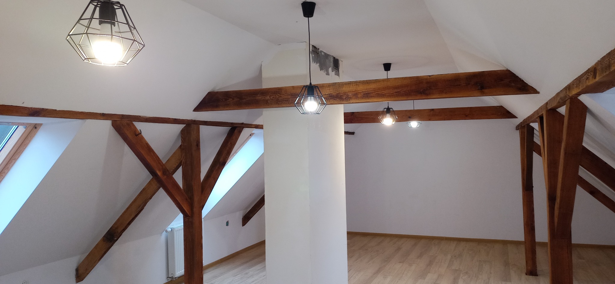 Wnętrze poddasza z drewnianą konstrukcją stropu, białymi ścianami, jasną podłogą i trzema lampami wiszącymi w metalowych kloszach o geometrycznym kształcie, widoczne ślady po remoncie na suficie.