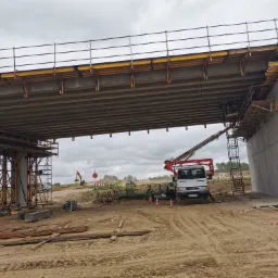 Budowa wiaduktu drogowego z widocznym podnośnikiem koszowym na platformie roboczej, szalunki, zbrojenia i elementy konstrukcyjne w trakcie montażu.