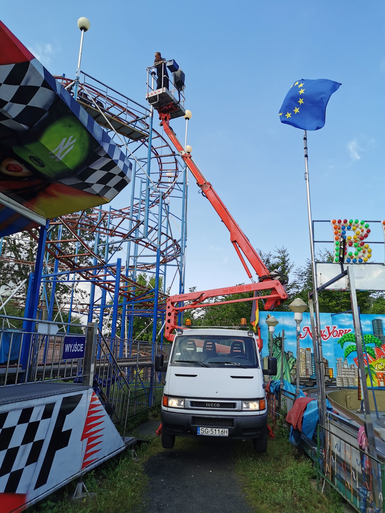 Pracownik w koszu podnośnika na wysokości, naprawia rollercoaster w wesołym miasteczku, flaga Unii Europejskiej w tle.