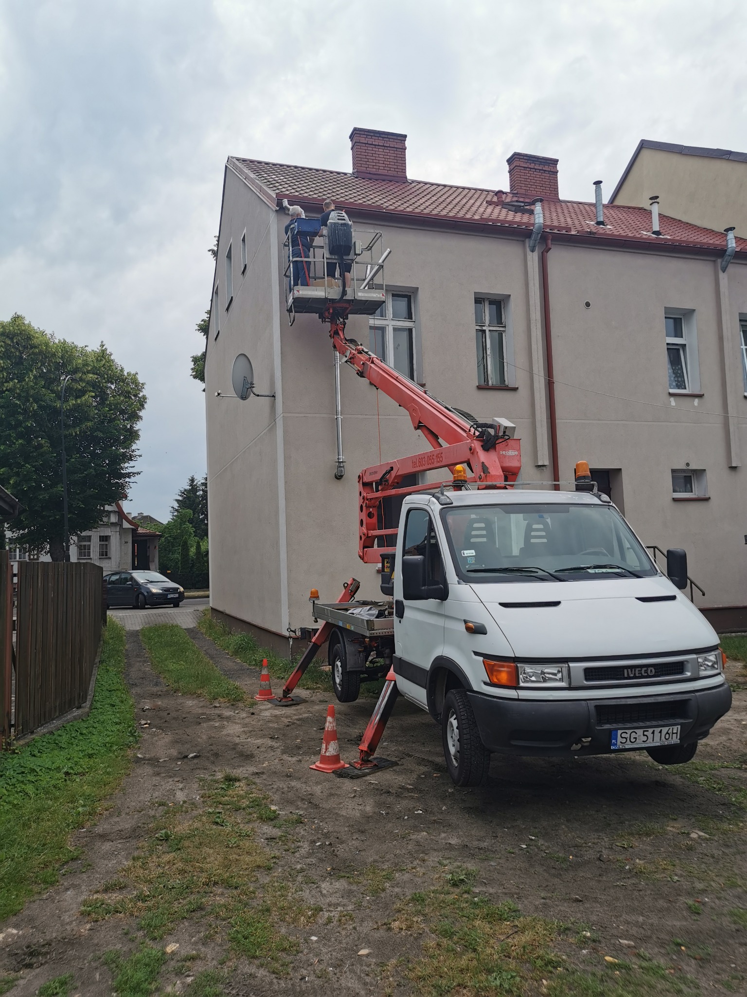 Dwóch pracowników w koszu podnośnika, przy elewacji budynku z czerwonym dachem, biały samochód Iveco z podnośnikiem koszowym na podwórku, zabezpieczony pomarańczowymi pachołkami.