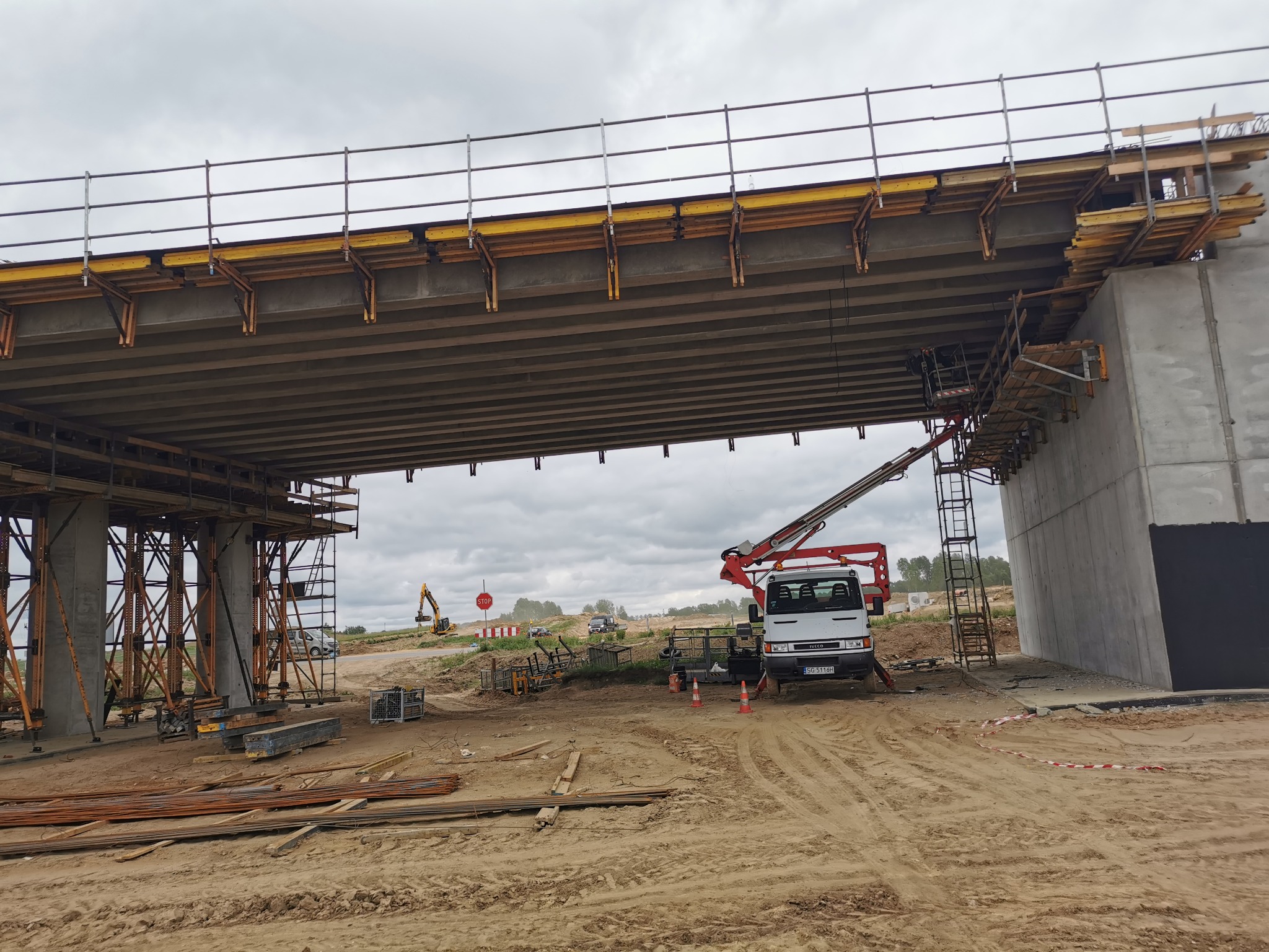Budowa wiaduktu drogowego z widocznym podnośnikiem koszowym na platformie roboczej, szalunki, zbrojenia i elementy konstrukcyjne w trakcie montażu.
