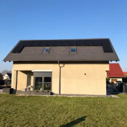 Dom jednorodzinny z panelami słonecznymi na dachu pokrytym szarą dachówką, widok z poziomu trawnika w słoneczny dzień.