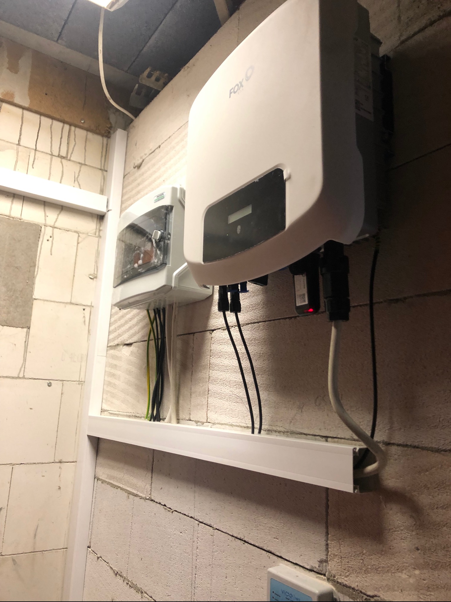 Biała obudowa falownika firmy FOX oraz skrzynka elektryczna zamontowane na ścianie z bloczków betonowych, widoczne przewody i elementy montażowe.