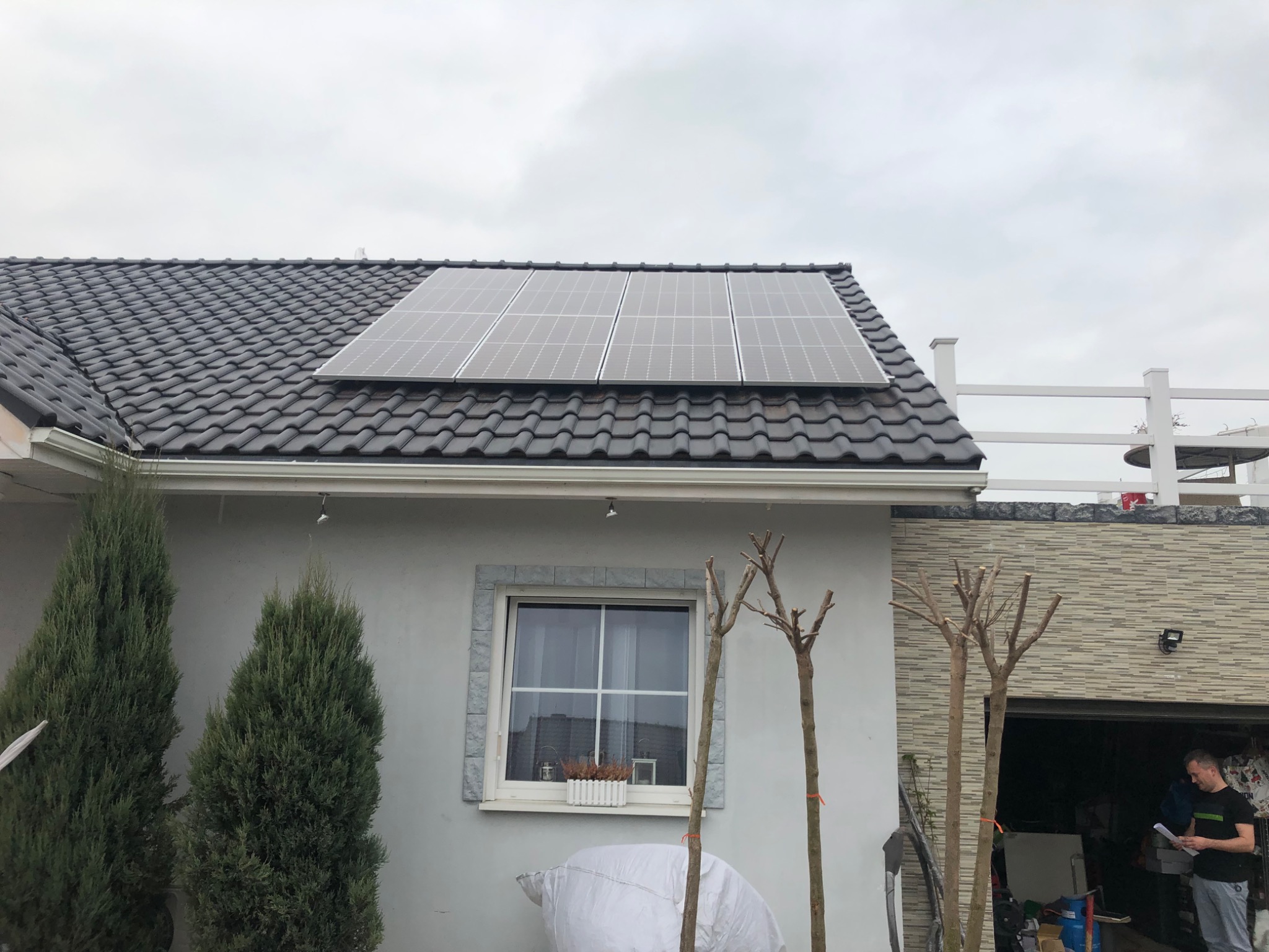 Fotowoltaika na dachu domu jednorodzinnego z ciemną dachówką, widok z poziomu ogrodu, z widocznymi iglakami i młodymi drzewkami przed elewacją.