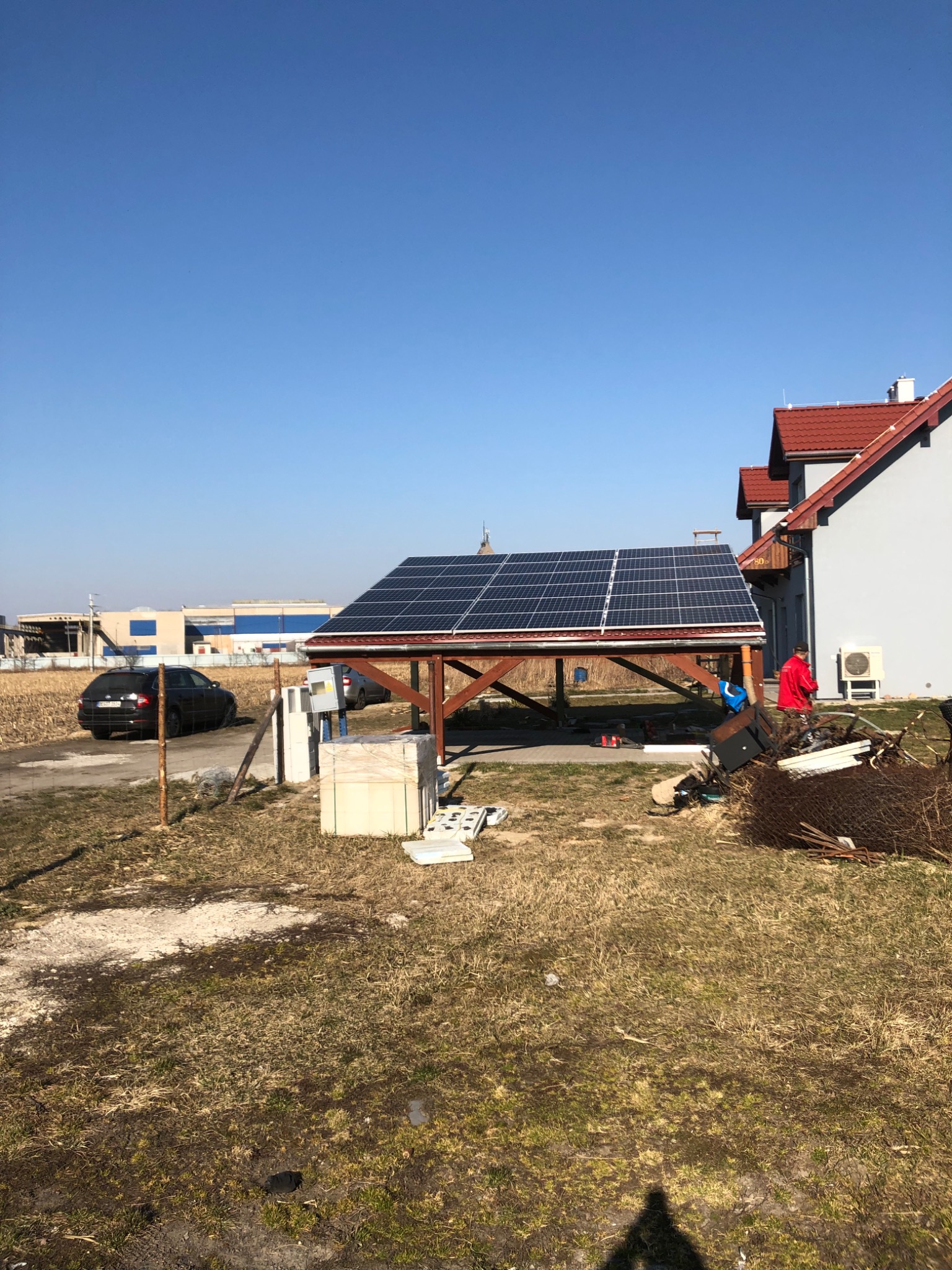 Drewniana wiata z panelami słonecznymi na tle domu jednorodzinnego i błękitnego nieba, widok z zewnątrz posesji, z widocznym samochodem i elementami ogrodzenia.