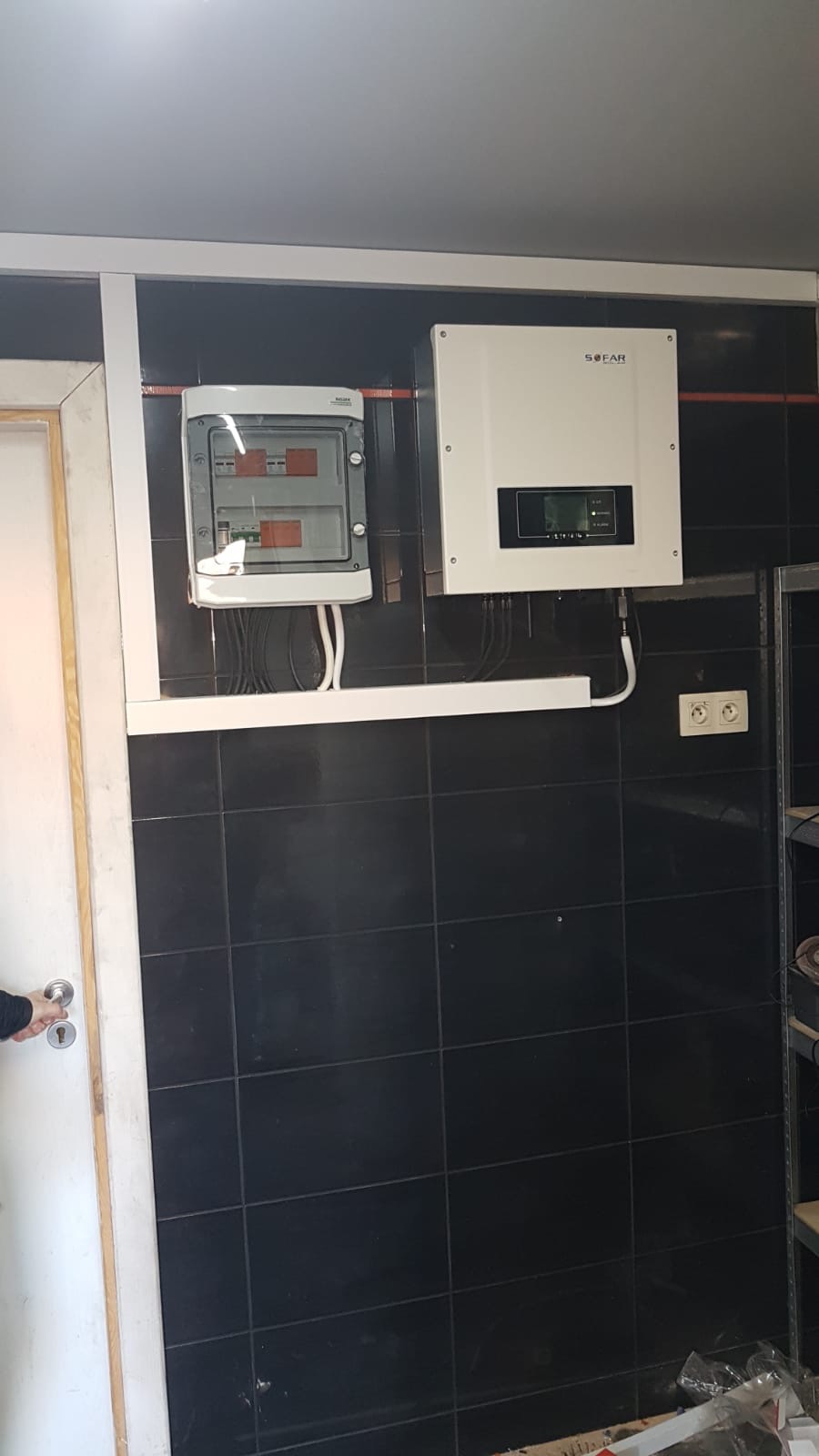 Instalacja elektryczna z białym inwerterem SOFAR i skrzynką rozdzielczą na czarnej, kafelkowanej ścianie w pomieszczeniu technicznym.