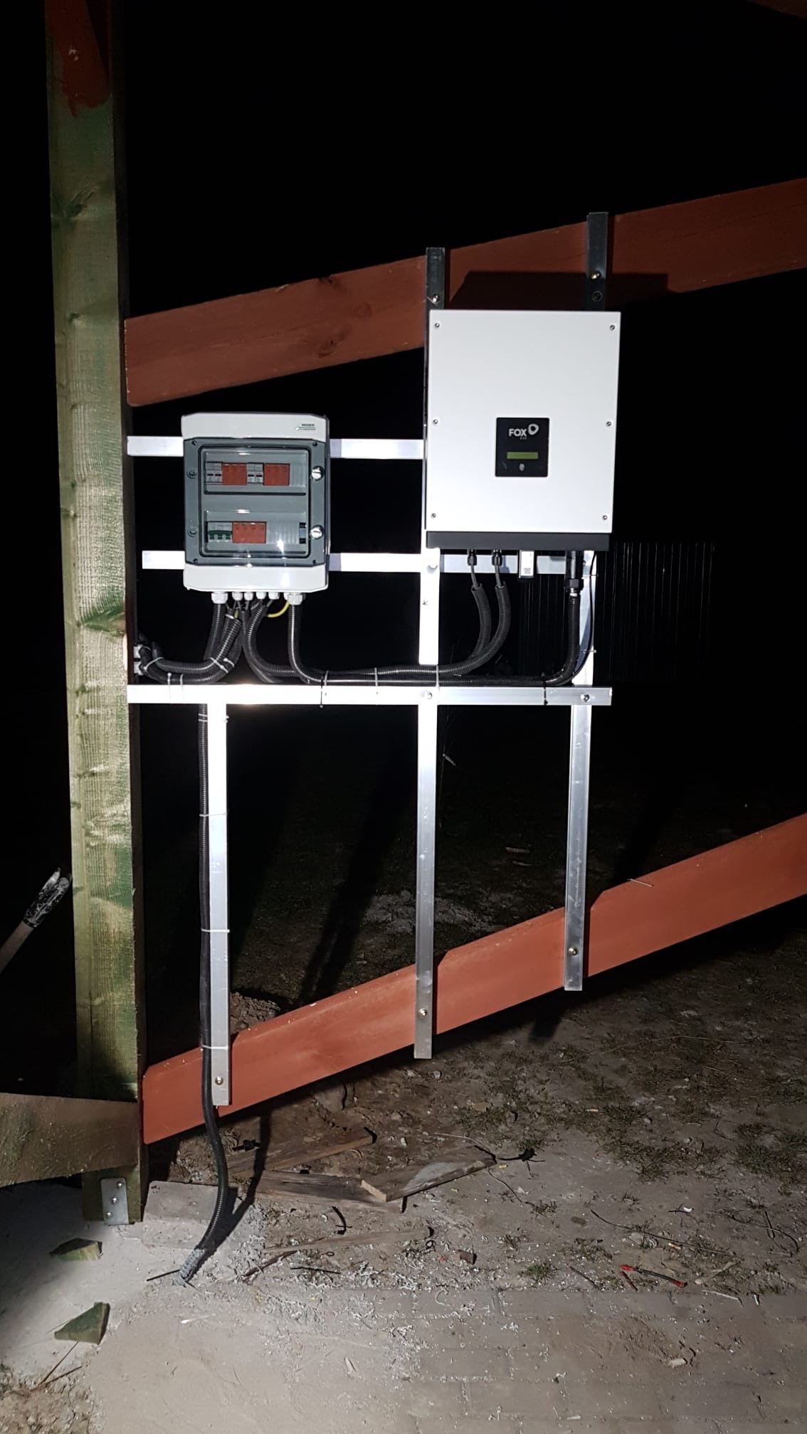 Zamontowany inwerter FOX i skrzynka elektryczna na konstrukcji z drewna, widoczne przewody i elementy montażowe w słabym oświetleniu.