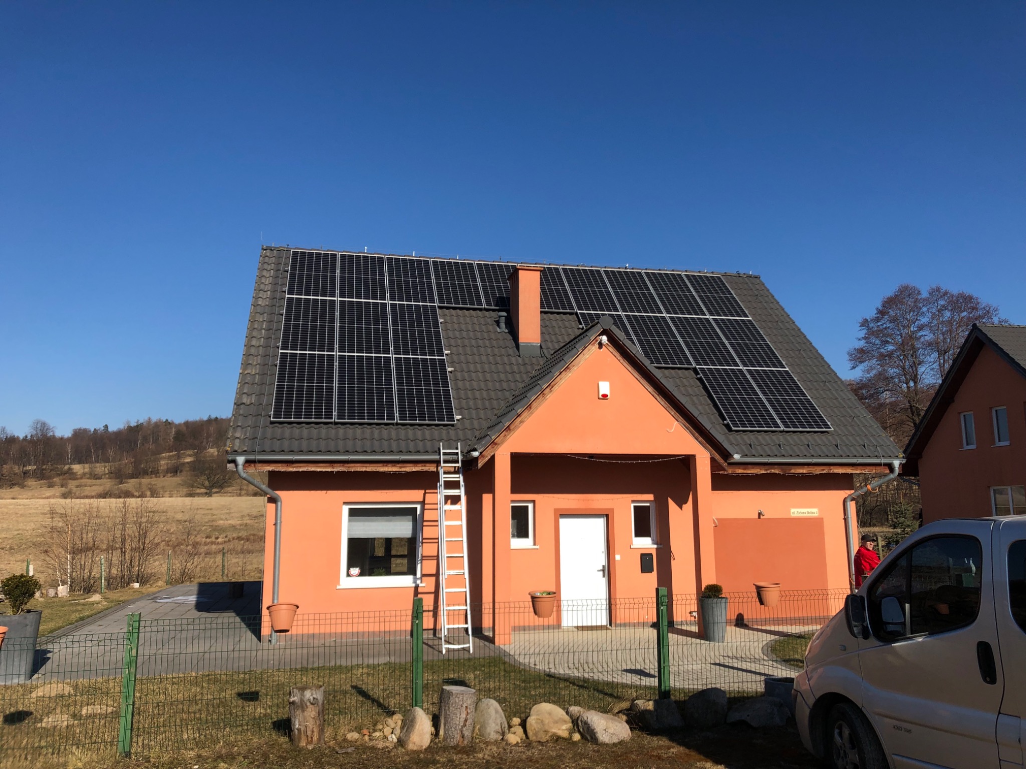 Pomarańczowy dom jednorodzinny z panelami słonecznymi na dachu, drabina oparta o ścianę, zaparkowany samochód dostawczy w pobliżu.