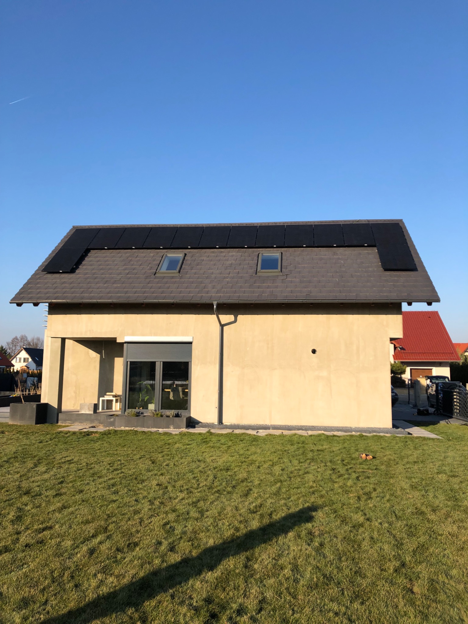 Dom jednorodzinny z panelami słonecznymi na dachu pokrytym szarą dachówką, widok z poziomu trawnika w słoneczny dzień.