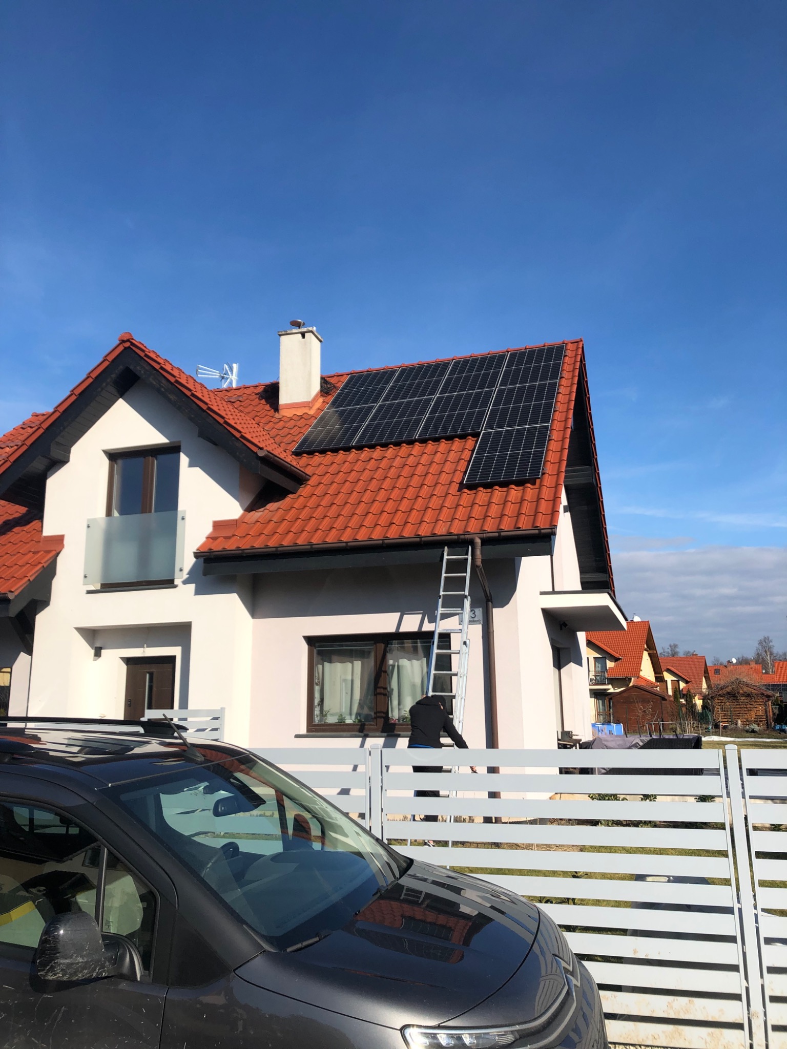 Montaż paneli fotowoltaicznych na dachu domu jednorodzinnego z czerwonej dachówki, widoczna drabina i fragment sylwetki montera na tle błękitnego nieba.