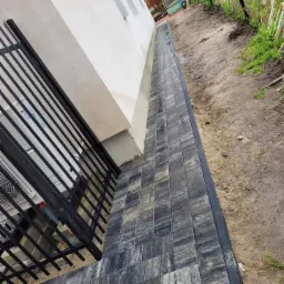 Ułożona kostka brukowa wzdłuż ściany budynku i metalowego ogrodzenia, widoczny fragment elewacji i niezagospodarowany teren.