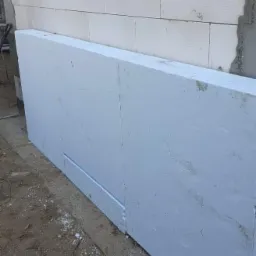 Płyta styropianowa przylegająca do ściany z białych bloczków, widoczne docięte krawędzie i drobne resztki materiałów budowlanych na ziemi.