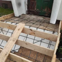 Przygotowanie do wylewki schodów wejściowych: drewniany szalunek z siatką zbrojeniową na styropianie, widoczne stare, zielone drzwi i fragment elewacji budynku.