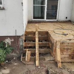 Przygotowanie do wylewki betonowej schodów zewnętrznych, widoczne drewniane szalunki, piasek, fragment elewacji i roślinności.