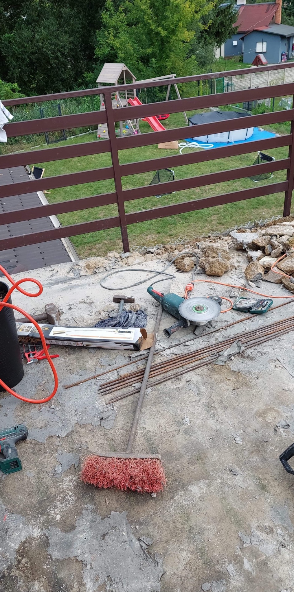 Przygotowania do prac budowlanych na tarasie: widoczne narzędzia, pręty zbrojeniowe, szlifierka kątowa, młotki, przewody, szczotka i gruz. W tle brązowe ogrodzenie z widokiem na ogród z placem...