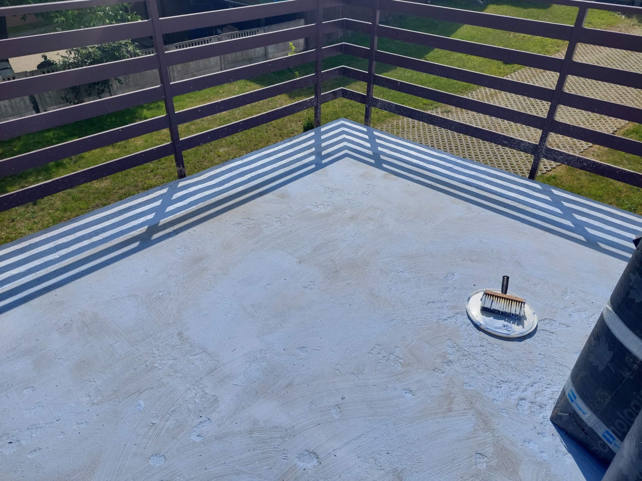 Szary taras w trakcie hydroizolacji, widoczny pędzel malarski na plastikowej tacy i rolka papy, w tle brązowe ogrodzenie i trawnik.