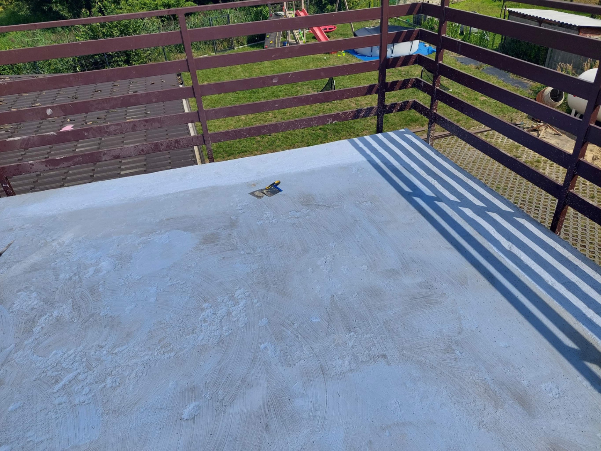 Świeżo wylana, szara, betonowa wylewka tarasu z widoczną pacą na powierzchni, otoczona brązową metalową barierką z widokiem na ogród z placem zabaw i betoniarką.