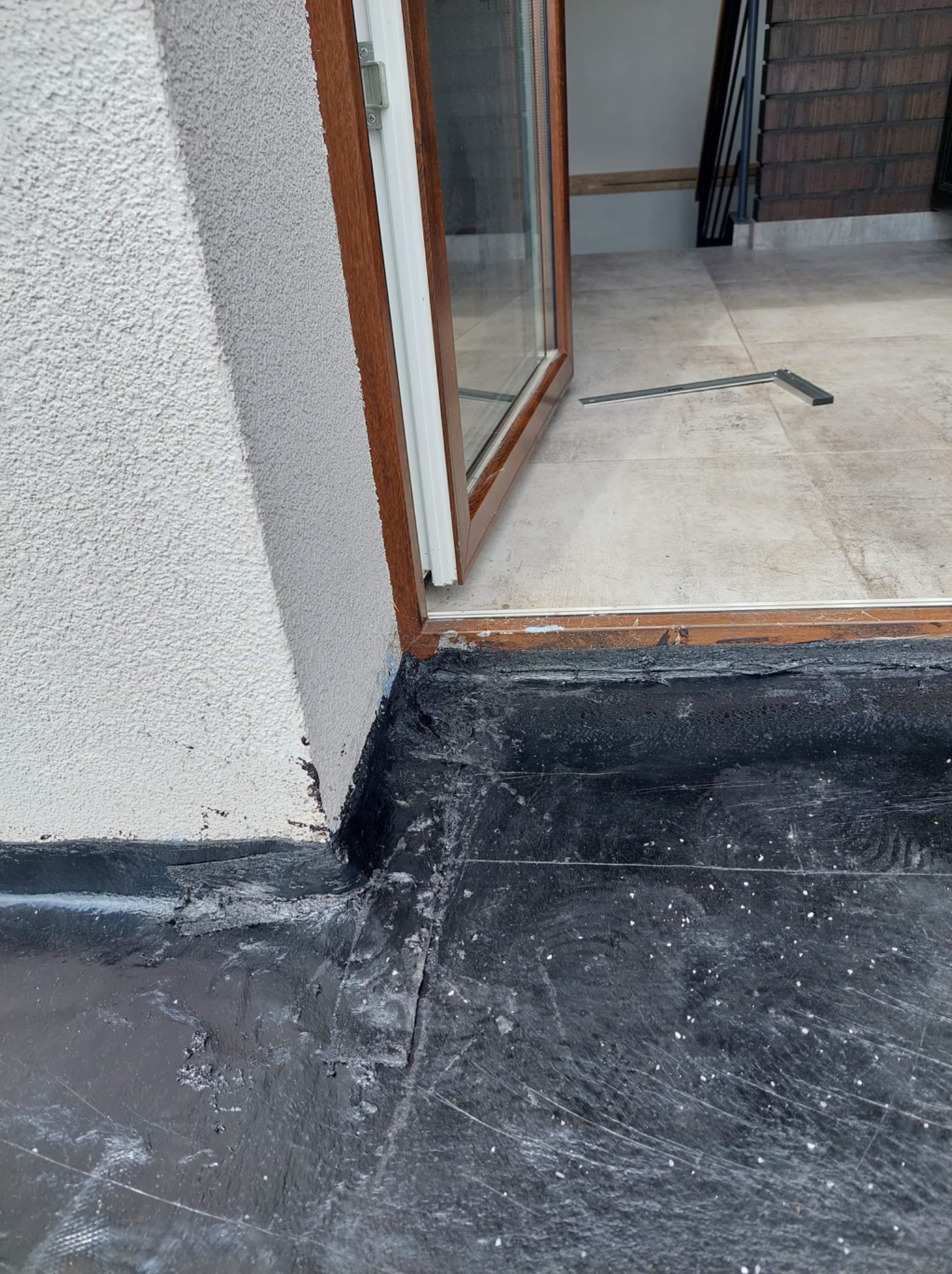 Fragment hydroizolacji przy progu drzwi balkonowych; widoczna warstwa bitumiczna łącząca się ze ścianą budynku pokrytą tynkiem strukturalnym oraz fragment posadzki balkonu z płytkami ceramicznymi.