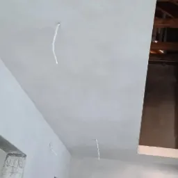 Świeżo otynkowany, jasnoszary sufit z wystającymi przewodami elektrycznymi, widok z dołu na otwartą przestrzeń stropu z drewnianą konstrukcją.