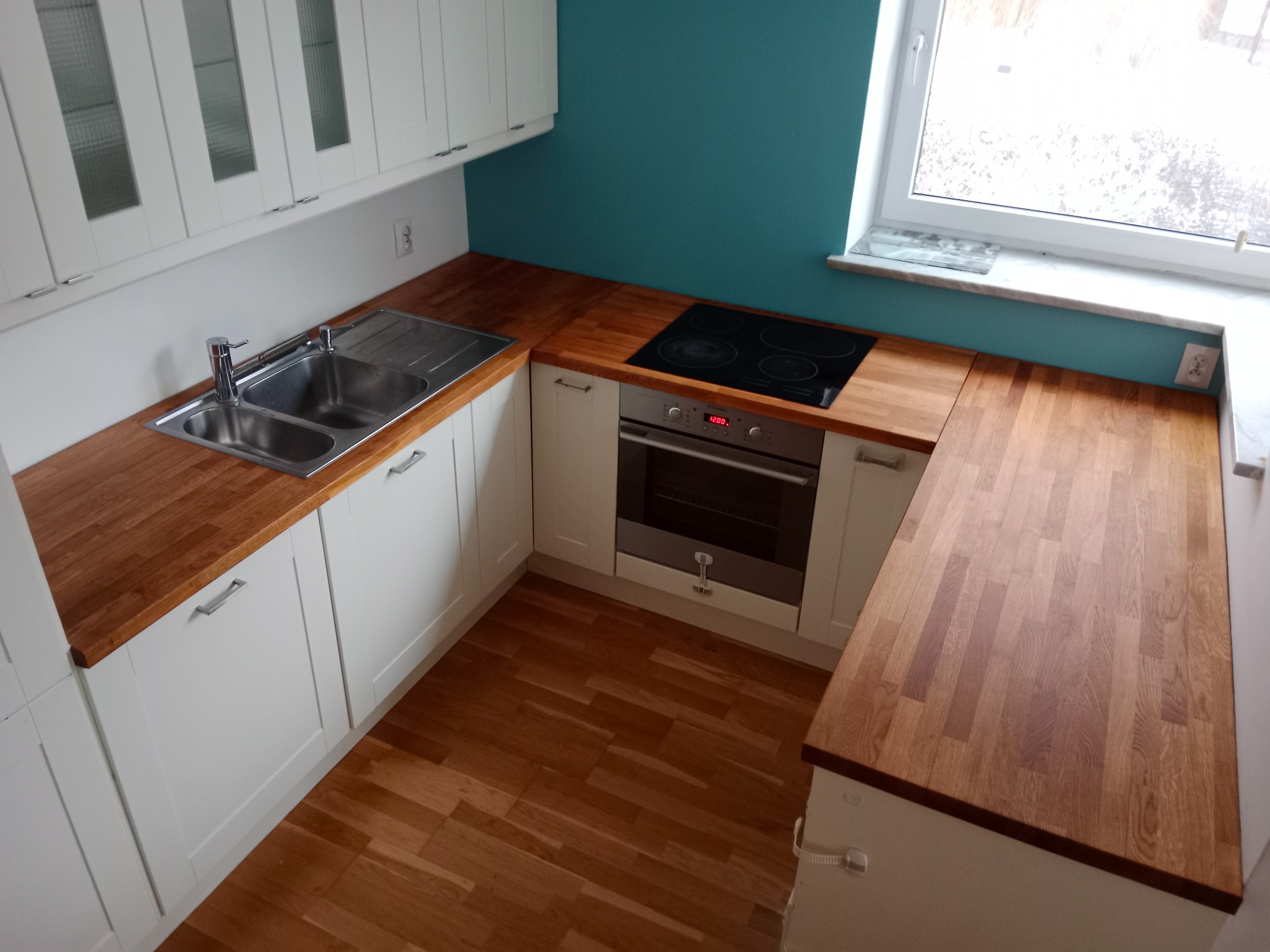 Wnętrze kuchni po remoncie: białe szafki z drewnianymi blatami, dwukomorowy zlewozmywak ze stali nierdzewnej, płyta indukcyjna, piekarnik, turkusowa ściana i drewniana podłoga w jodełkę.
