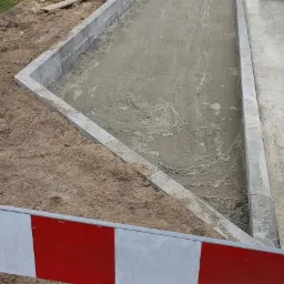 Przygotowanie podłoża pod nawierzchnię drogi: wyrównany piasek i obrzeża betonowe, widoczne narzędzia i maszyny budowlane w tle.