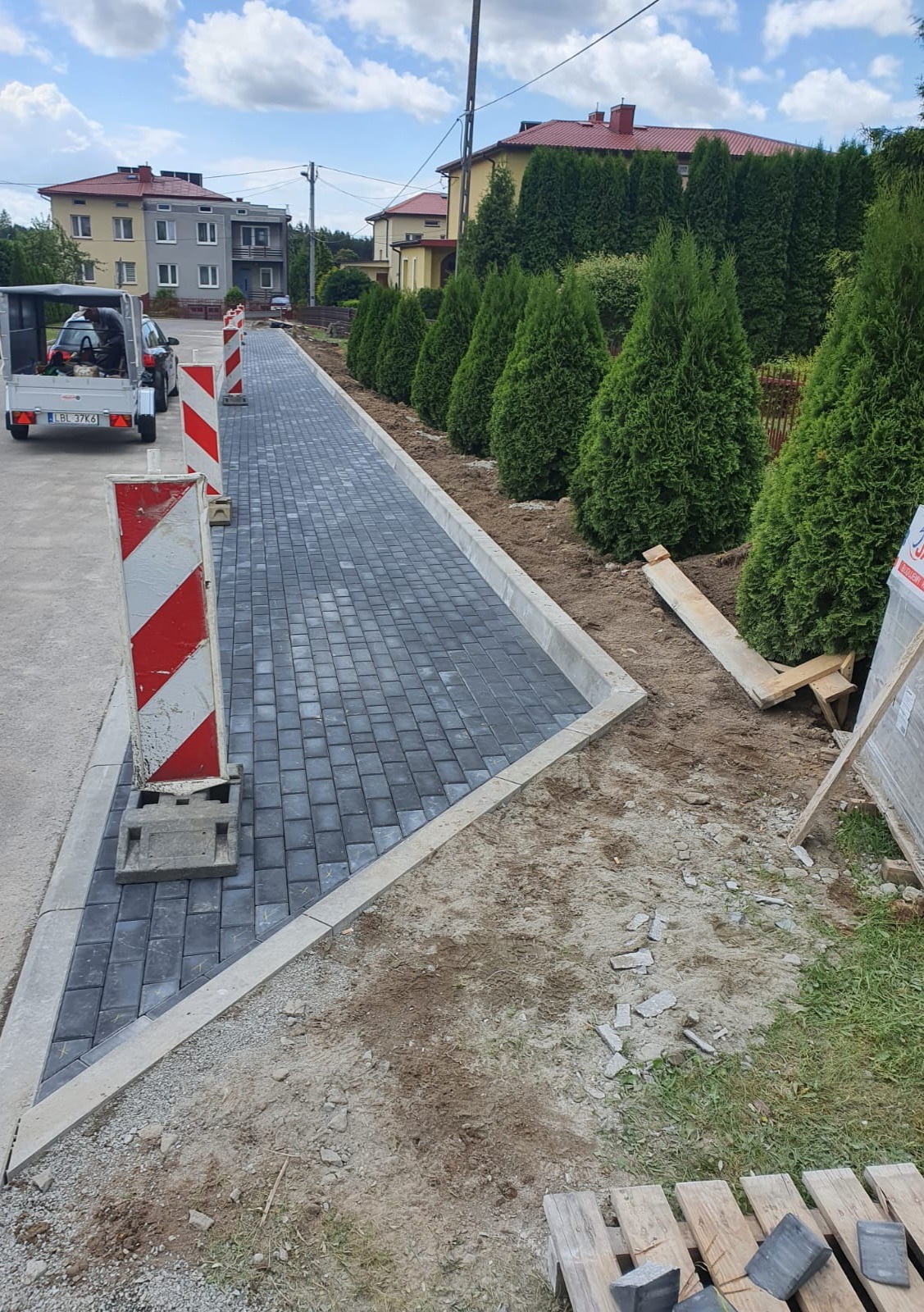 Nowo ułożony chodnik z szarej kostki brukowej, obrzeża betonowe, widoczne zabezpieczenia budowlane w postaci słupków z czerwono-białymi pasami, tło: budynki mieszkalne i rząd tuj.