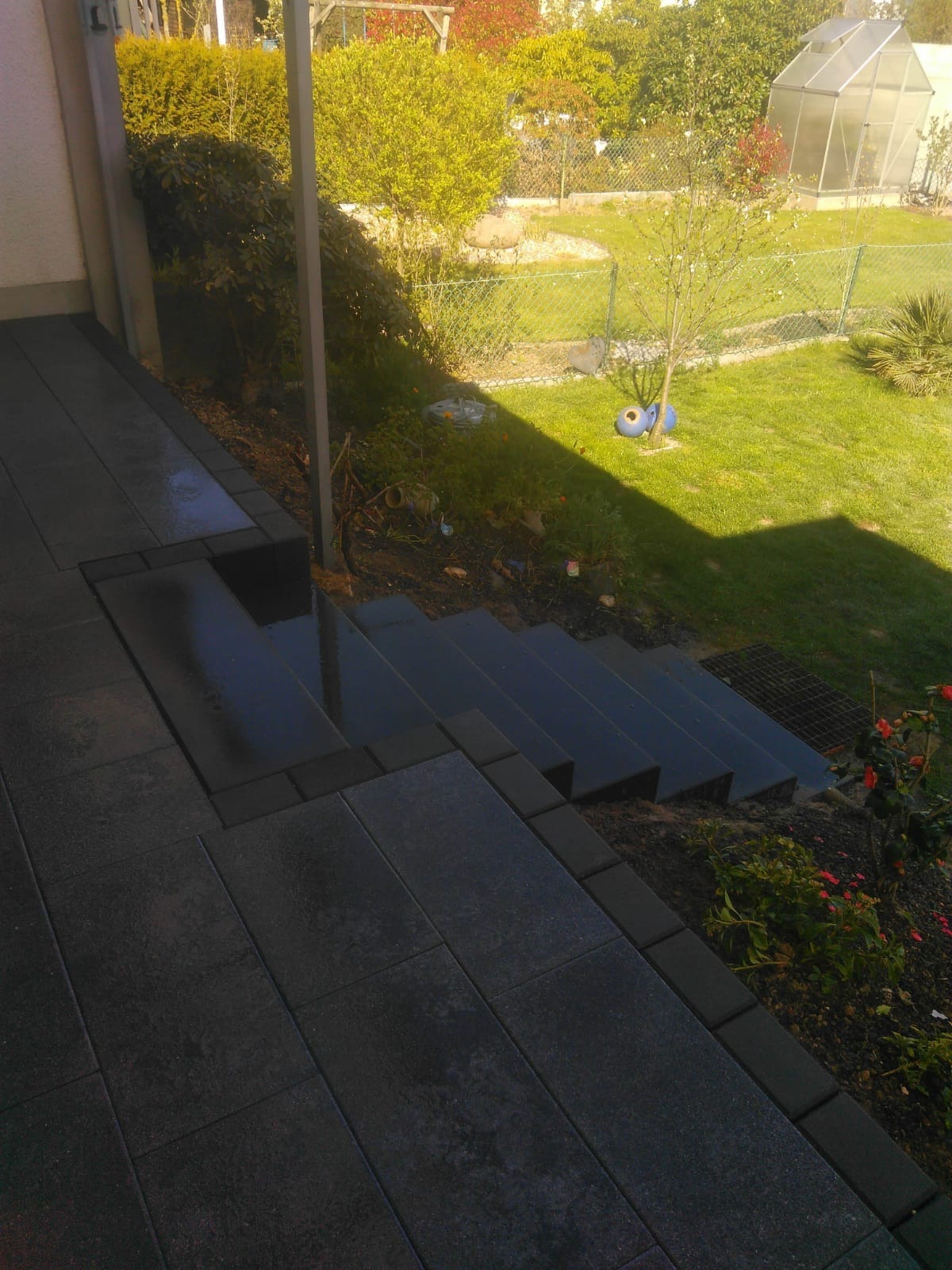 Ciemnoszare, prostokątne płyty chodnikowe ułożone jako ścieżka i schody prowadzące w dół ogrodu, z widoczną szklarnią w tle i odblaskami wody na stopniach.