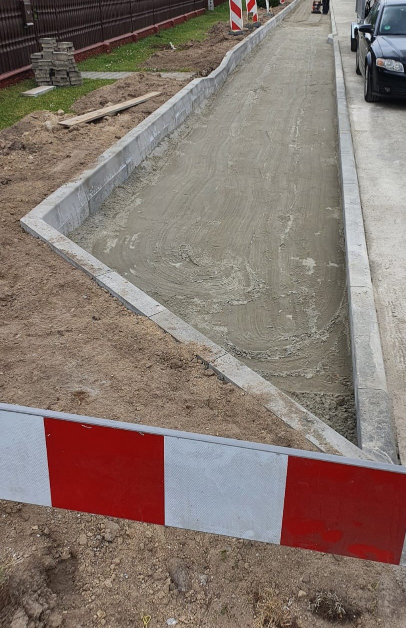 Przygotowanie podłoża pod nawierzchnię drogi: wyrównany piasek i obrzeża betonowe, widoczne narzędzia i maszyny budowlane w tle.