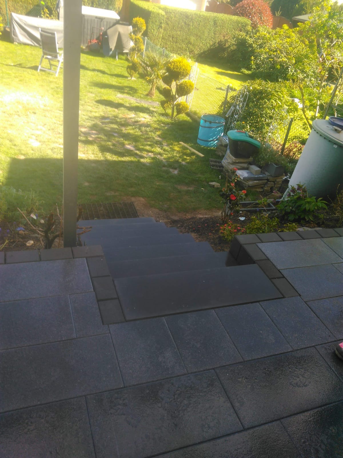 Schody wyłożone ciemną kostką brukową prowadzące do ogrodu z zielonym trawnikiem, widoczne elementy architektury ogrodowej w tle.