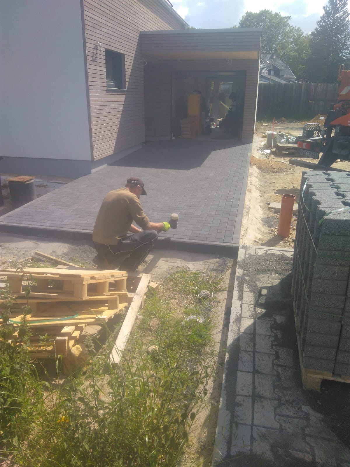 Pracownik w trakcie wbijania obrzeża kostki brukowej młotkiem gumowym, widoczna ułożona nawierzchnia podjazdu, paleta z kostką, w tle dom z garażem