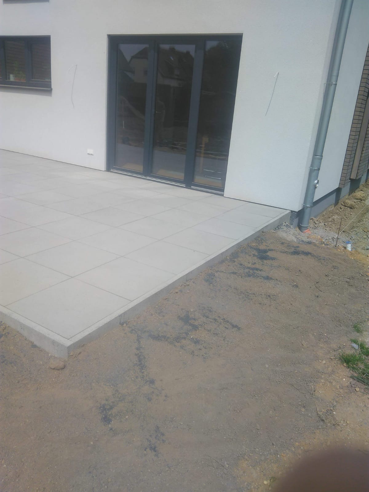 Jasnoszara, kwadratowa płyta tarasowa ułożona przy ścianie budynku z szarymi drzwiami balkonowymi, widoczny fragment rynny i przygotowany teren pod dalsze prace ziemne.