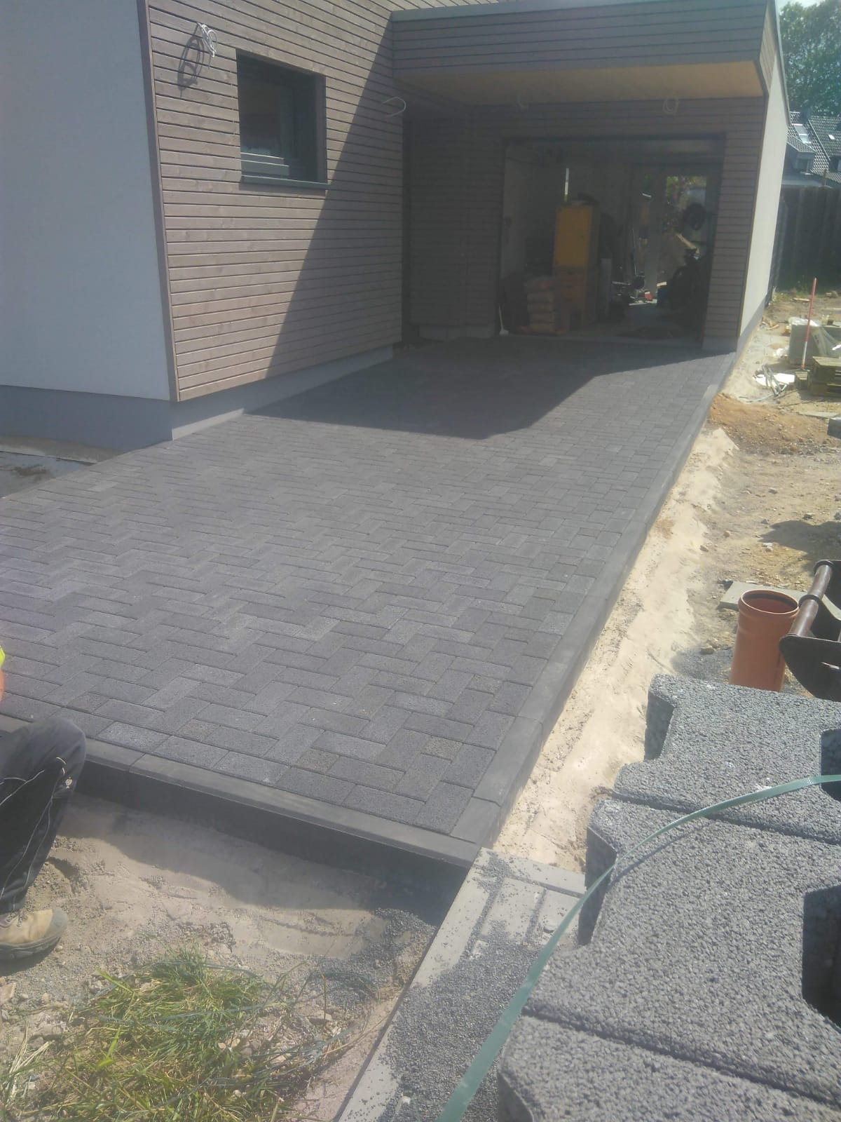 Ułożona, szara kostka brukowa w jodełkę prowadząca do garażu z drewnianą elewacją. Widoczne obrzeża, piasek i stos kostki brukowej czekającej na ułożenie.