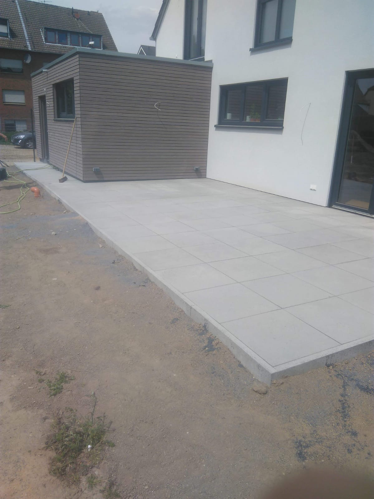 Nowo ułożony, jasnoszary taras z kwadratowych płyt betonowych przy nowoczesnym domu z drewnianą dobudówką, widoczny fragment ogrodu w trakcie prac wykończeniowych, z pozostawioną miotłą opartą...