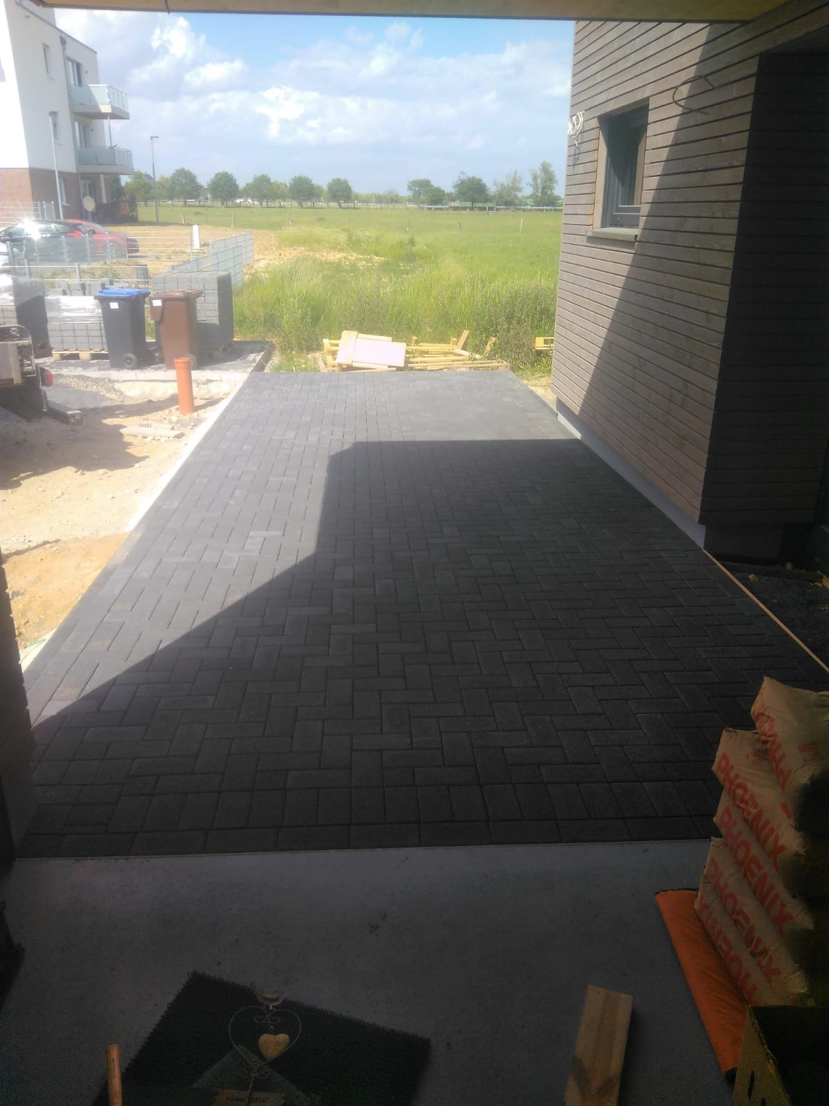 Ułożona, ciemnoszara kostka brukowa na podjeździe domu z drewnianą elewacją, widok z zadaszonego tarasu, w tle trawnik i budynek mieszkalny.